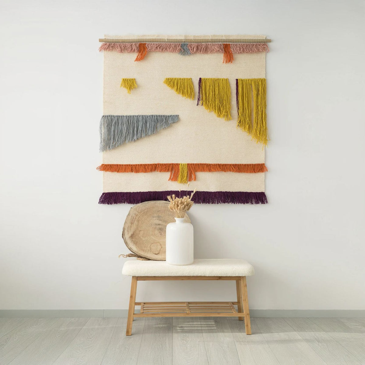 Handwoven tapestry Gelo with vibrant geometric patterns and colorful fringes, crafted from wool and hung on a bamboo rod, displayed in a modern interior with natural accents.
